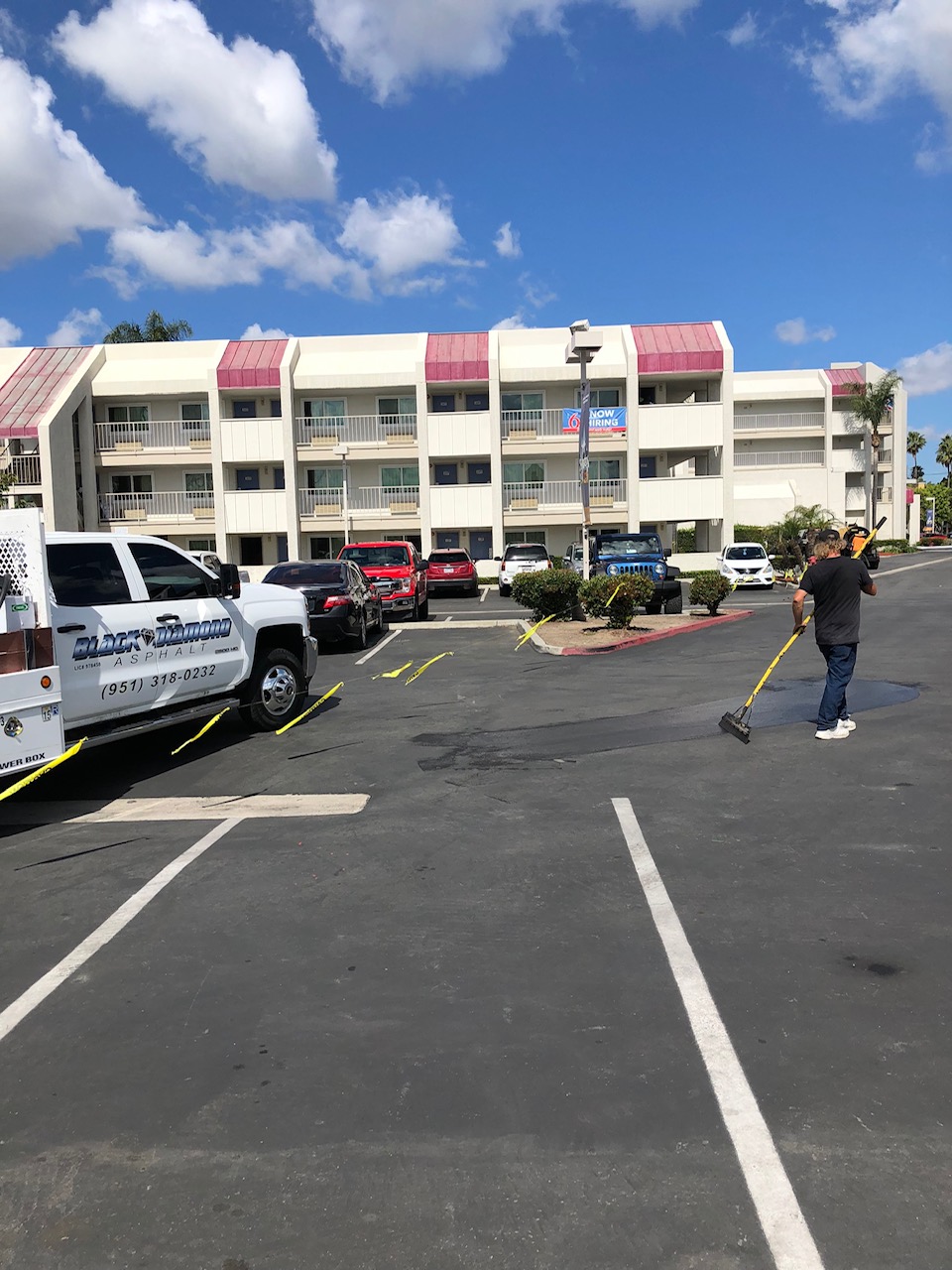 A man is patching the road in front of a building, with a Black Diamond Asphalt van parked along with other cars